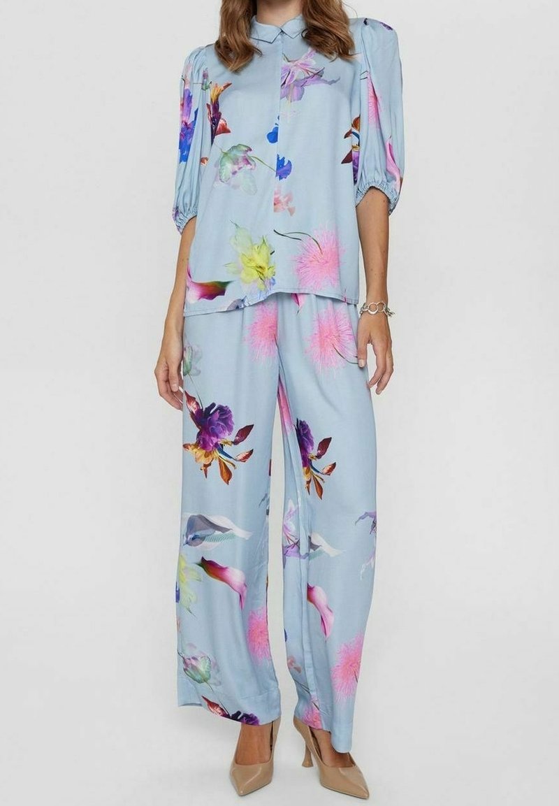 Femme portant un chemisier bleu clair à motif floral avec des manches bouffantes et un pantalon large assorti, associé à des talons hauts beiges et un bracelet.
