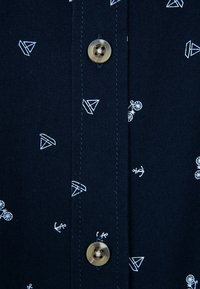 Navy skjorta med knappar som har vita geometriska mönster och cykelmönster, med en rund knappdetalj och kontrasterande sömmar. Mjuk tygtextur.