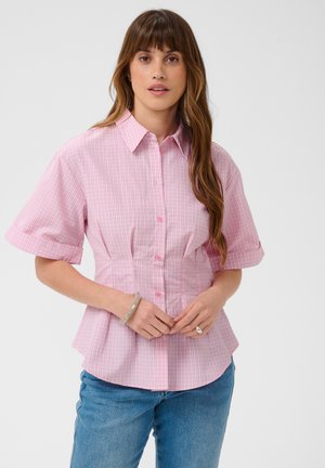 Femme aux longs cheveux bruns portant une chemise rose à manches courtes boutonnée et un jean bleu, debout devant un fond clair uni.