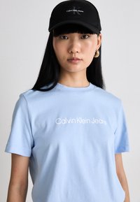 Světle modré bavlněné tričko s bílým logem "Calvin Klein Jeans", doplněné černou kšiltovkou se stejným logem. Hladká textura, design s kulatým výstřihem.
