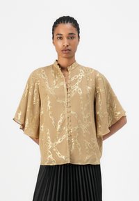 Blusa di kain emas muda dengan pola rantai, lengan pendek bergetar, kerah berdiri, dan desain depan berbutang.