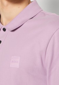 Polo léger violet clair en tissu texturé, avec un col classique et trois boutons noirs, portant un petit logo "BOSS" en relief.