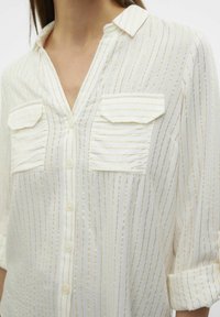 Chemise blanche à boutons avec de fines rayures verticales dorées. Comprend deux poches poitrine, un col et des manches retroussées. Tissu léger.