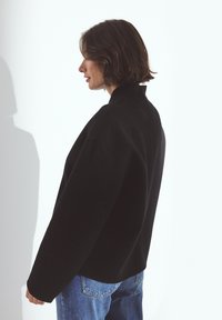 Woman with short brown hair wears an oversized black jacket and blue jeans, standing sideways against a white background with shadow.