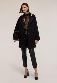 Abrigo de lana negro con cuello de muesca, con detalles de botones amarillos. Debajo, una blusa de encaje negro transparente, combinada con pantalones de cuero negro ajustados y tacones.