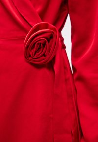 Czerwona tkaninowa odzież z dużym akcentem w kształcie róży w talii, o gładkiej fakturze i związanym pasku do zapięcia.