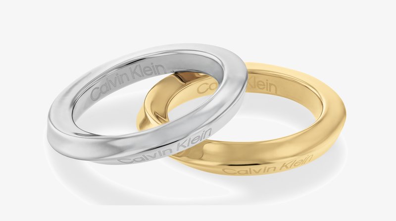 Two interlocking rings, one in polished silver and the other in gold. Both are smooth with engraved "Calvin Klein" on the inner side.