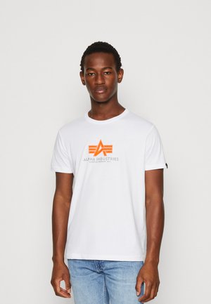 Hvid bomuld t-shirt med et grafisk design i orange og grå med teksten "ALPHA INDUSTRIES" nedenunder, rund hals og opfoldede ærmer.