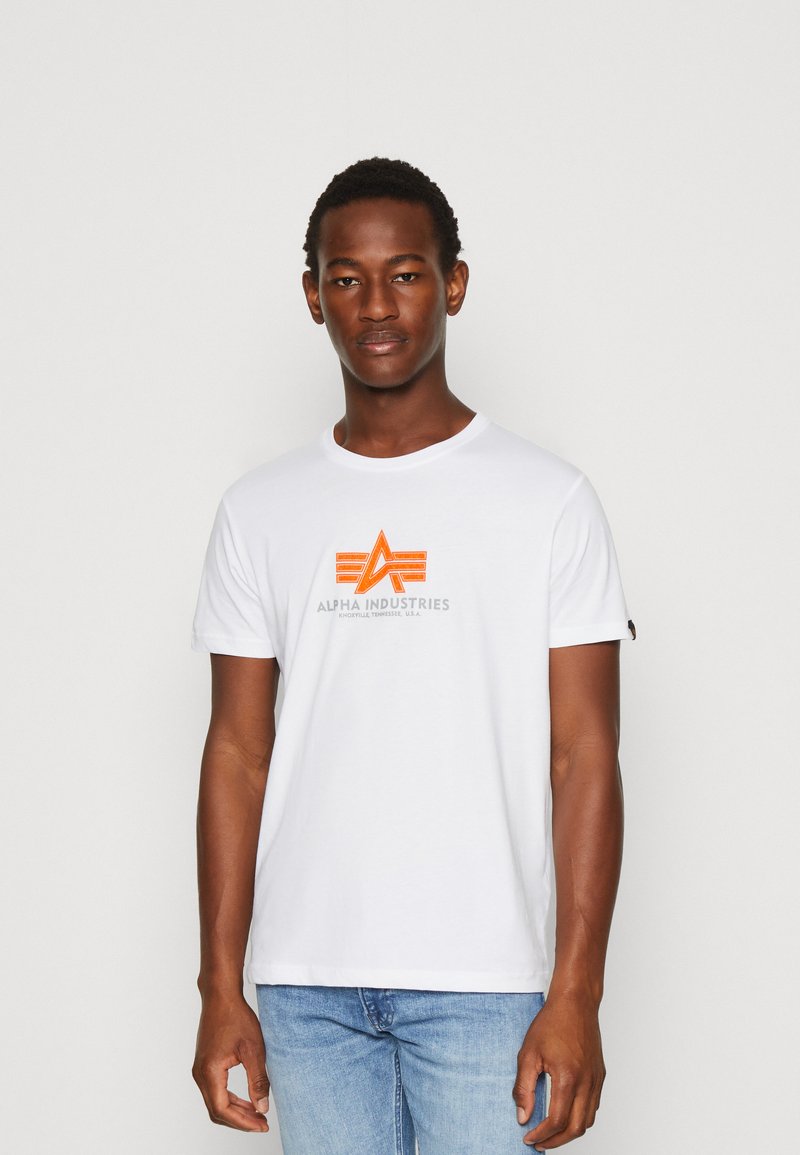 Baltos medvilninės marškinėliai su oranžine ir pilka grafika, po kuria užrašas "ALPHA INDUSTRIES", apvaliu iškirpimu ir sulenktais rankovėmis.