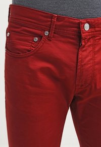Jeans in cotone rosso con vestibilità slim, caratterizzati da componenti in metallo argentato, cinque tasche e dettagli di cucitura in vista. Texture liscia con una leggera lucentezza.