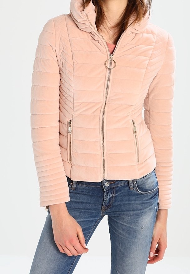 Femme portant une veste matelassée rose clair avec une tirette de fermeture éclair ronde, associée à un jean en denim bleu, sur un fond blanc uni.