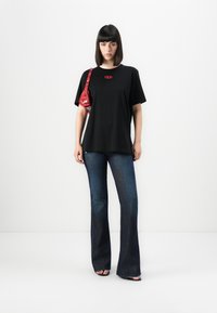 Black cotton t-shirt with small red logo, paired with flared blue denim jeans. The model holds a small red patterned bag.