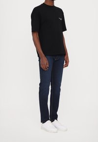 Černé tričko s krátkým rukávem a kulatým výstřihem, v kombinaci s tmavě modrými džíny a bílými teniskami. Jednoduchý design bez viditelných vzorů.