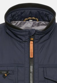 Veste matelassée bleu marine avec col montant, fermeture éclair avec détail en cuir et poche zippée sur le côté gauche. Texture lisse.