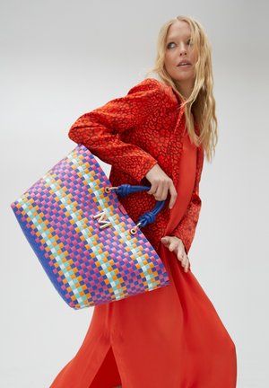 Vrouw in rode jurk en patroonjas met een grote kleurrijke gevlochten tas met blauwe handvatten en gouden "M" detail.