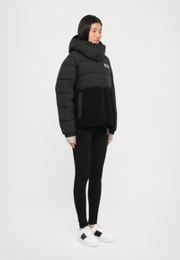 Zwarte pufferjack met hoge kraag, voorzien van een contrasterend zwart texturenpaneel en een frontale rits, gedragen over zwarte leggings en witte sneakers.