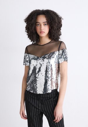 Kort topje met zilveren pailletten die een schitterend patroon creëren, voorzien van een zwarte doorzichtige mesh halslijn en korte mouwen. Gestreepte broek wordt eronder gedragen.