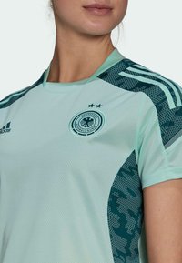 Osoba v světlé zelené dresu německého národního fotbalového týmu s černým orlem na hrudi a dvěma hvězdami nad ním.