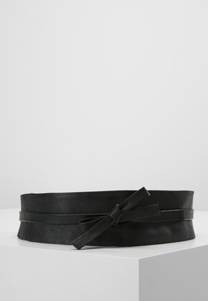 Ceinture large en cuir noir avec une lanière étroite nouée en nœud sur une surface blanche sur un fond gris uni.