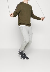 Felpa verde oliva con logo, pantaloni felpati grigi e sneakers nere. La felpa presenta dettagli a fantasia sulle maniche.