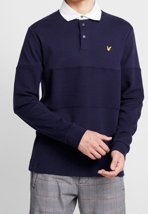 Homme portant un maillot de rugby bleu marine à manches longues avec un col blanc et un logo d'oiseau jaune, associé à un pantalon gris à carreaux.