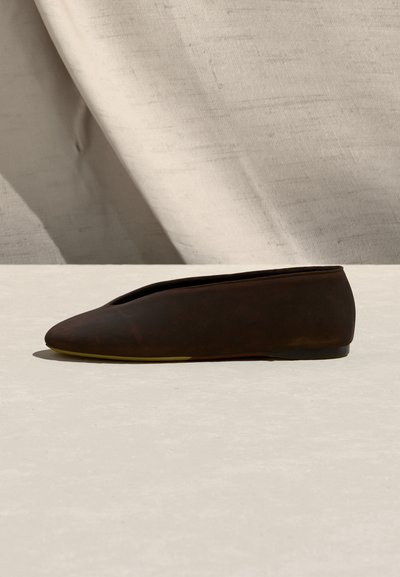 Chaussure plate marron foncé à bout pointu et talon bas, présentée sur une surface beige avec un fond en tissu clair.