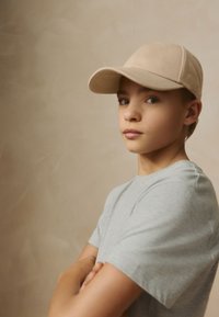 Cappellino beige con visiera curva, indossato sopra capelli castano chiaro. Maglietta grigia a maniche corte, braccia incrociate, su uno sfondo morbido e neutro.