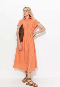 Robe chemise orange à rayures avec des manches courtes et une fermeture à boutons, assortie à un sac frangé marron et des sandales à lanières marron.