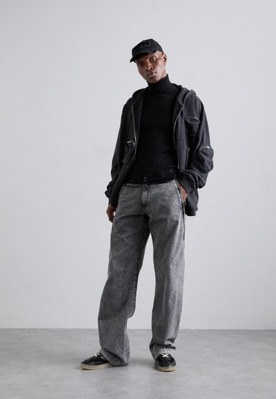 Μαύρο φούτερ με φερμουάρ πάνω από μαύρο ψηλό γιακά, γκρι φαρδιά τζιν και μαύρα αθλητικά παπούτσια. Το καπέλο είναι μαύρο με ανάγλυφη λεπτομέρεια λογότυπου.