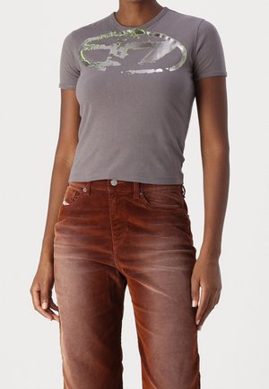 Vrouw draagt een aansluitend grijs t-shirt met korte mouwen met metalen afbeelding en een hoog taille roestkleurige corduroy broek.