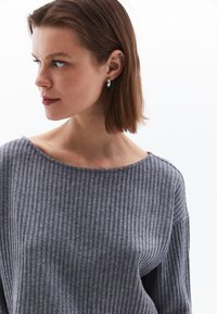 Grijze geribbelde trui met een wijd decollet&eacute; en verlaagde schouders, gecombineerd met zilveren oorbellen met een ring, op een effen witte achtergrond.