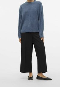 Person som bär en lös blåtröja, vida svarta cropped byxor och svarta ballerina-skor mot en enhetlig vit bakgrund.