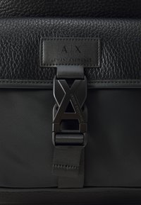 Sac à dos noir avec une partie supérieure en cuir texturé, une partie inférieure en nylon lisse et une boucle noire angulaire avec des détails du logo "AX".