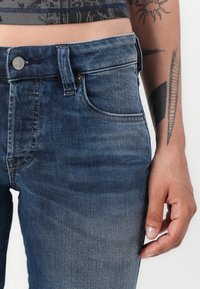 Tumši zili džinsi ar klasisko piecu kabatu dizainu, aprīkoti ar rāvējslēdzēju un metāla pogu aizdari. Gluda tekstūra un neliela izbalēšana.