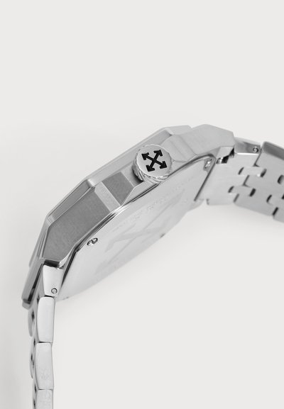 Gros plan sur une montre-bracelet argentée, avec son boîtier anguleux, son bracelet métallique et sa couronne ornée d’un symbole noir représentant des flèches croisées.