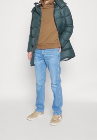 Chaqueta acolchada verde con capucha, sudadera marrón, vaqueros de mezclilla azul claro y zapatillas beige con detalles blancos. Materiales suaves y corte casual.