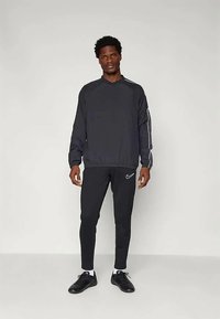 Muž stojící v černém dlouhém rukávu sportovním topu, černých sportovních kalhotách Nike s logem, bílých ponožkách a černých sportovních botách na jednobarevném pozadí.