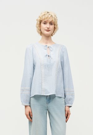 Νέος με κοντά σγουρά ξανθά μαλλιά που φοράει ανοιχτό μπλε μπλούζα με μακριά μανίκια και λεπτομέρειες δαντέλας, καθώς και ανοιχτόχρωμα τζιν, στέκεται μπροστά σε λευκό φόντο.