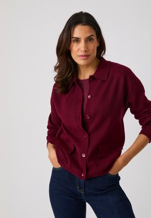 Donna con capelli scuri che indossa un cardigan rosso scuro abbottonato e jeans blu scuro, in piedi con le mani nelle tasche davanti a uno sfondo semplice.