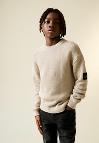 Bézs színű kötött pulóver texturált mintával, kerek nyakkivágással és bordázott mandzsettákkal. Sötétszürke nadrággal viselve. Fekete címke a bal ujján.