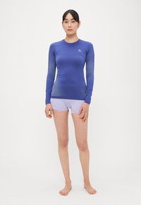 Μπλούζα με μακριά μανίκια σε μπλε χρώμα με αντίθετη υφή από δίχτυ, συνδυασμένη με ανοιχτόμωβ σορτς. Το υλικό φαίνεται ελαστικό και εφαρμοστό.