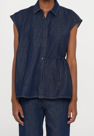 Dunkelblaue ärmellose Jeansbluse mit Knöpfen vorne und seitlicher Schleife, getragen mit passender dunkelblauer Jeanshose.