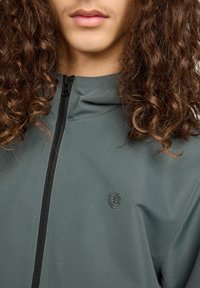 Person mit lockigem braunem Haar, die eine graue Zip-Jacke mit einem kleinen runden Logo auf der Brust trägt. Die Lippen sind sichtbar, aber keine anderen Gesichtszüge werden gezeigt.