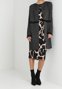 Žena v dlouhém šedém texturovaném kabátu přes černé a béžové midi šaty s abstraktním vzorem a černých kotníkových botách, stojící před bílou zdí.