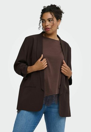 Jeune femme aux cheveux bouclés portant des boucles d'oreilles créoles, un blazer marron foncé, une chemise marron et un jean bleu tenant les revers du blazer.