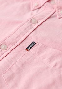 Rosa skjorta med knäppning gjord av texturerat tyg, med en knäppt krage, pärlliknande knappar och en framficka med en "SUPERDRY"-etikett.