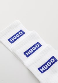 Drei Paar weiße Baumwollsocken mit einem blauen gewebten "HUGO"-Logo an den Bündchen, die eine gerippte Textur und ein klares Design aufweisen.