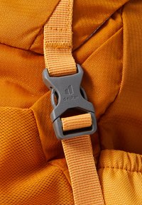 Orange fabric backpack strap with a gray plastic buckle. Textured surface with visible stitching and a logo on the buckle.