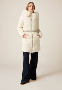 Vit vadderad jacka med huva av fuskpäls, silverdetalj vid midjan, dragkedja och sidofickor. Modellen bär mörka utsvängda byxor.