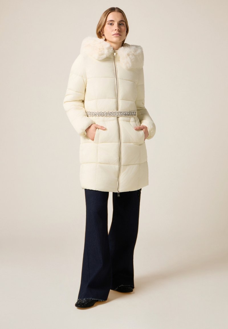 Vit vadderad jacka med huva av fuskpäls, silverdetalj vid midjan, dragkedja och sidofickor. Modellen bär mörka utsvängda byxor.